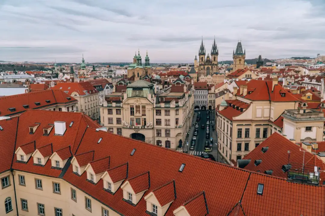Widok na dachy Pragi, z widocznymi wieżami kościołów i ulicą z samochodami, gdzie porusza się komunikacja miejska.