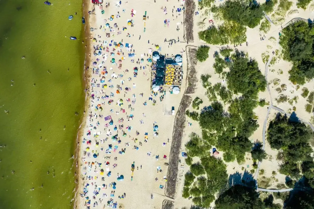 Tłum ludzi na piaszczystej plaży, kąpielisko i zielone drzewa. To widok z lotu ptaka, który pokazuje, że Litwa ma dostęp do morza.