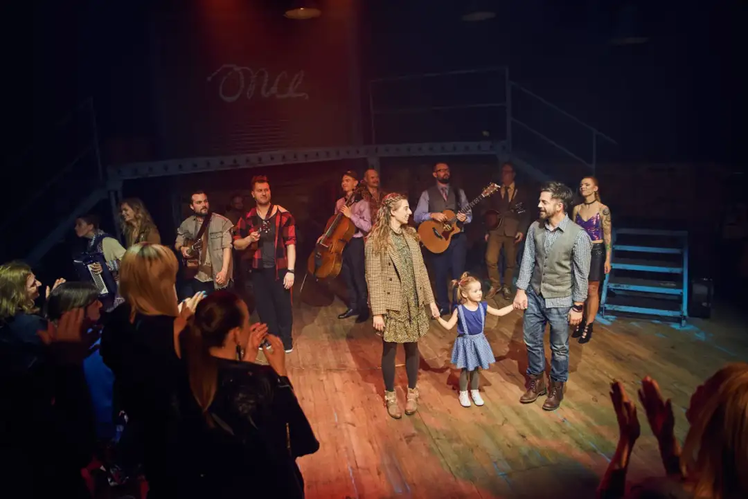 Aktorzy i muzycy z spektaklu "Once" na scenie, z małą dziewczynką trzymającą za rękę.