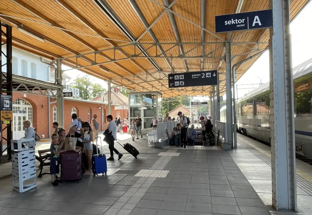 Ludzie z bagażami czekają na peronie. Znak "sektor A" i "peron 2" sugerują, że to stacja kolejowa, gdzie można dowiedzieć się, jak dojechać do Kołobrzegu pociągiem.