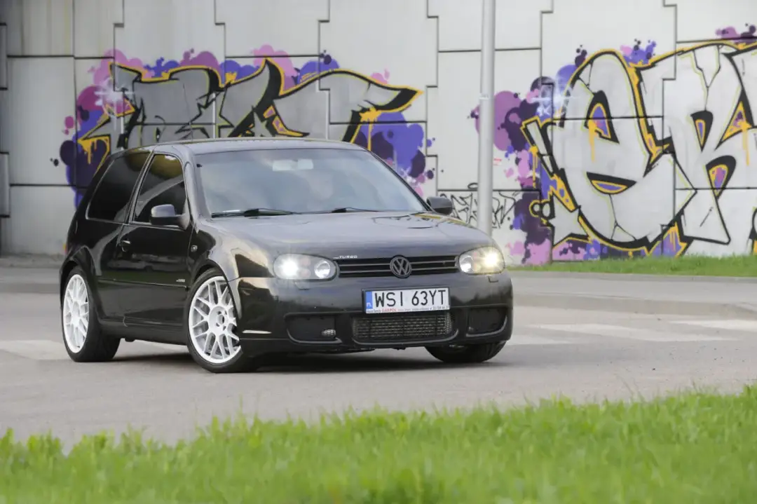Czarny Golf 4 z tuningowym body kitem i białymi felgami, zaparkowany na tle kolorowego graffiti.