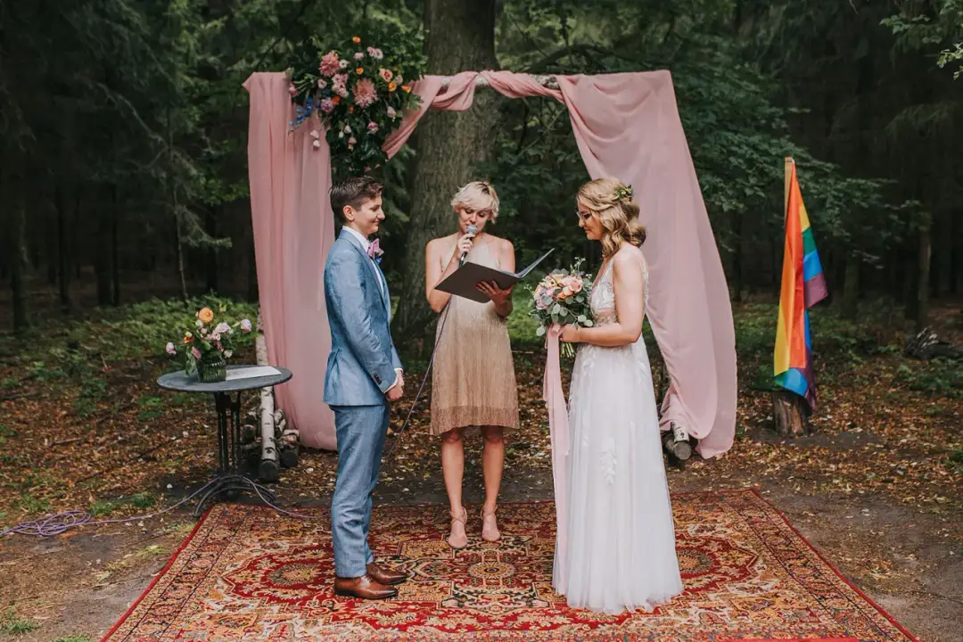 Ceremonia ślubna w lesie, gdzie można wziąć ślub homoseksualny. Dwie kobiety, jedna w garniturze, druga w sukni ślubnej, stoją przed urzędniczką. Tęczowa flaga symbolizuje miłość.