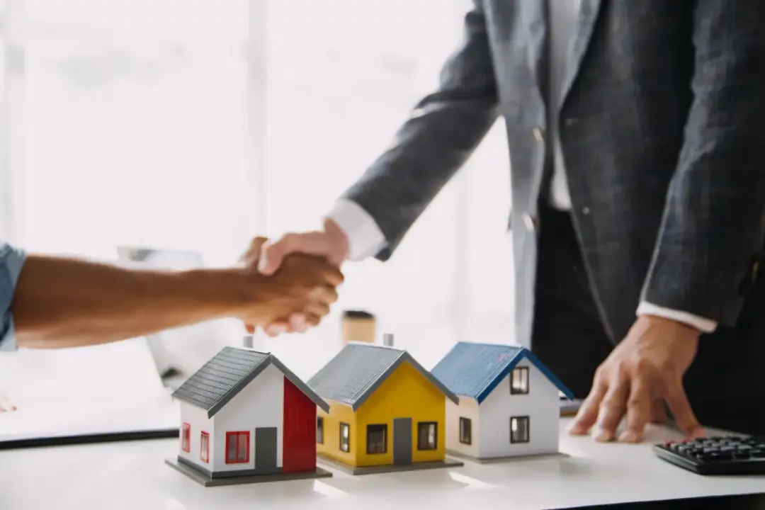 Uścisk dłoni symbolizuje udaną zamianę domu na mieszkanie. Trzy modele domów na stole.