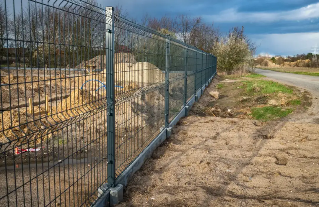 Trwa montaż ogrodzenia panelowego. Nowy płot z betonową podmurówką oddziela plac budowy od drogi i terenów zielonych.