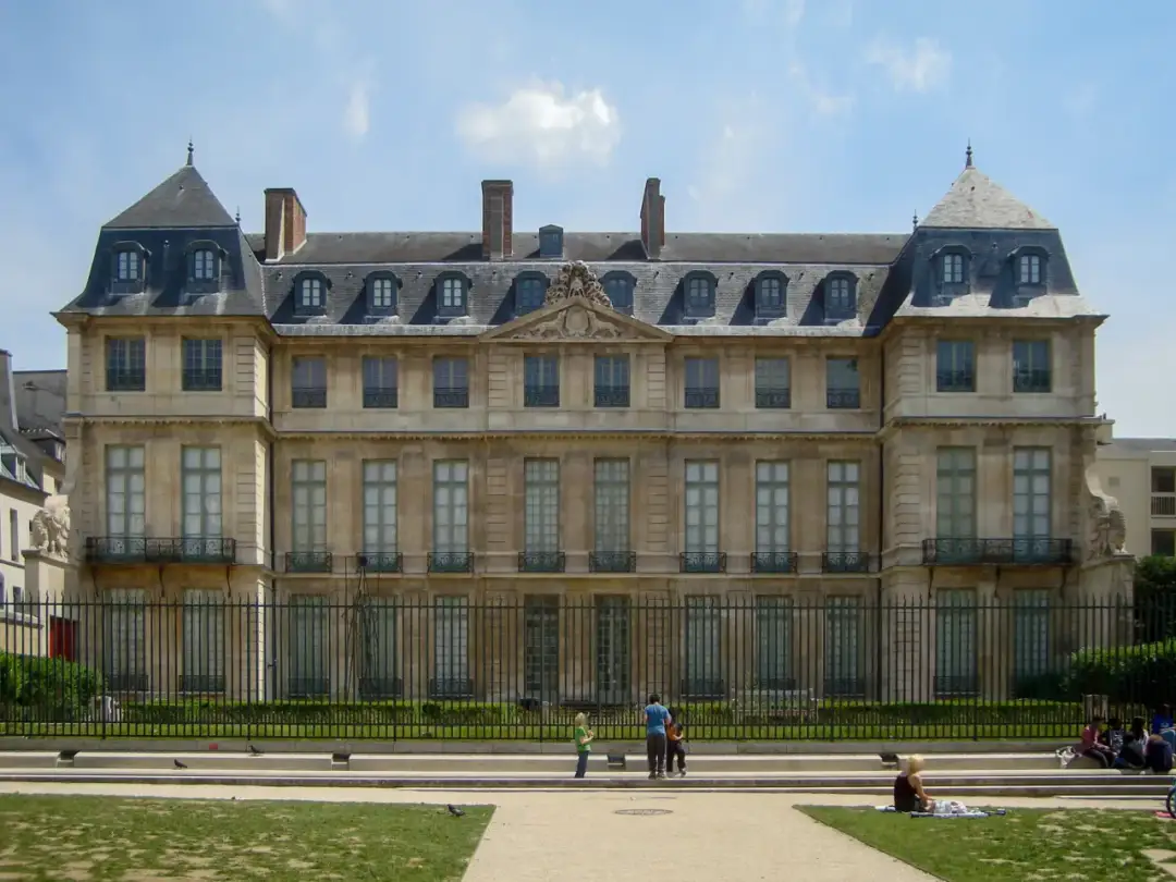 Elegancki budynek muzeum Picassa w Paryżu, z charakterystycznym dachem i oknami. W parku przed nim odpoczywają ludzie.