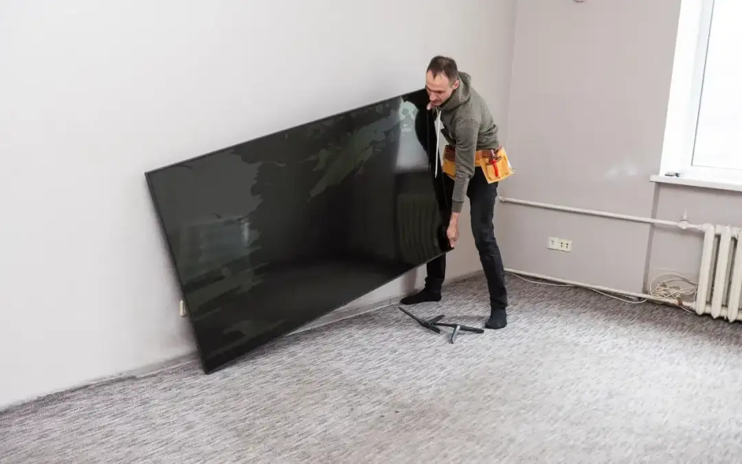 Mężczyzna z pasem narzędziowym przymierza się do powieszenia dużego telewizora. Zastanawia się, jak wysoko powiesić TV, by był idealnie widoczny.