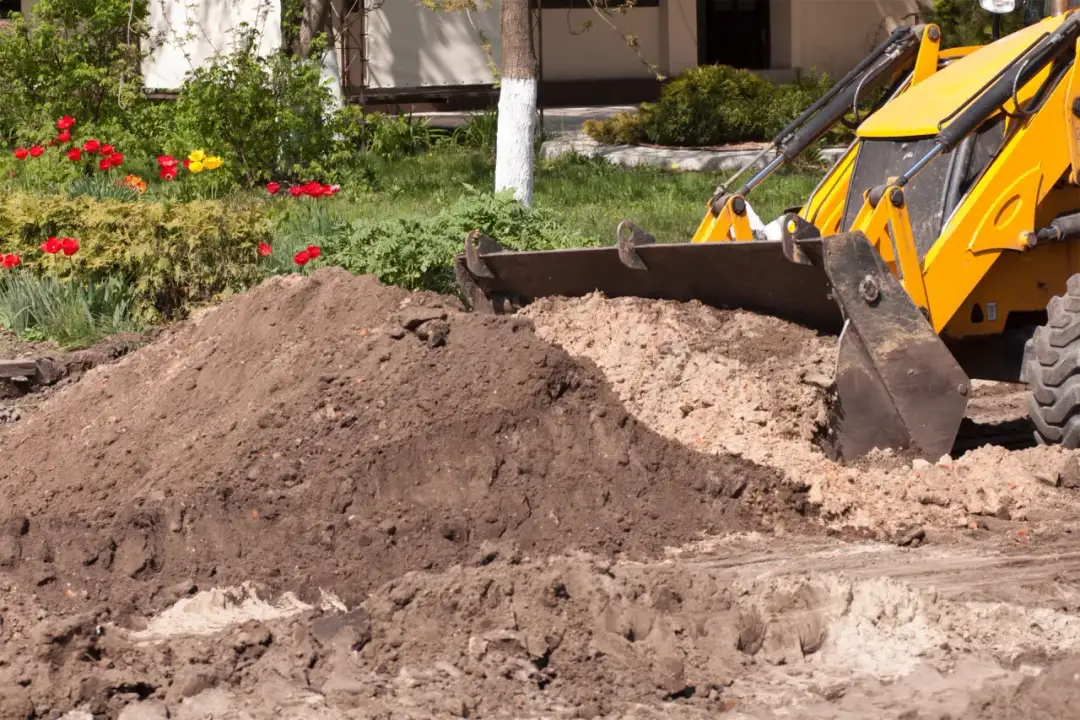 Koparka spycha ziemię, tworząc kopiec. W tle kwitnące tulipany i zielone krzewy. To jak rejestr wniosków decyzji i zgłoszeń, gdzie każda łopata to nowy etap.