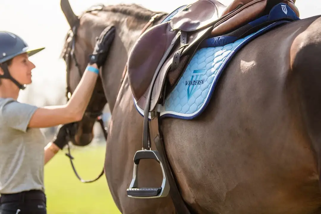 Jeździec głaszcze konia. Na grzbiecie zwierzęcia widać siodło i jasnoniebieski czaprak z logo.