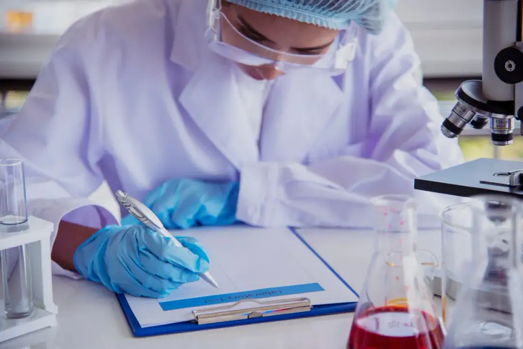 Osoba w fartuchu laboratoryjnym i rękawiczkach pisze na kartce obok mikroskopu i probówek.