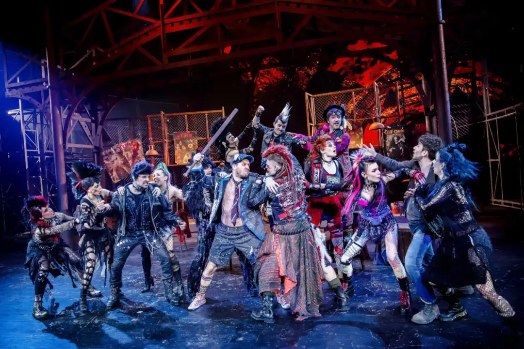 Grupa punkowych postaci na scenie, w tym z irokezami i w skórzanych kurtkach, w dynamicznej scenie z musicalu "We Will Rock You".