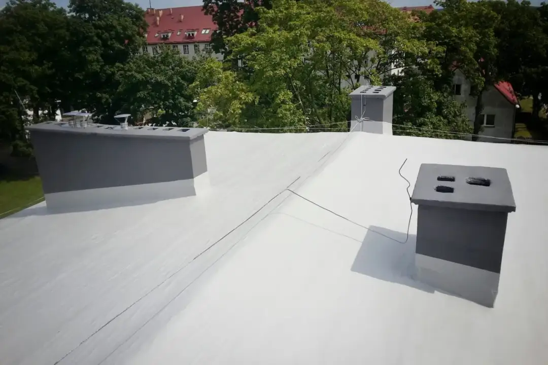 Nowy dach z białą hydroizolacją na papę, z szarymi kominami i zielenią w tle.