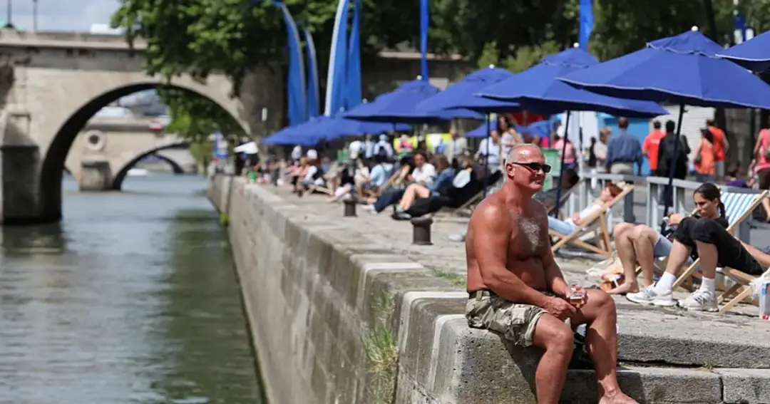 Ludzie odpoczywają nad Sekwaną, pod niebieskimi parasolami, w cieniu mostu.