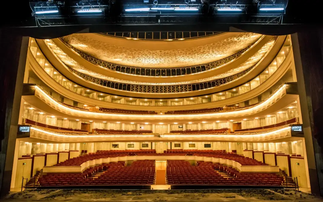 Wielki teatr, plan widowni z rzędami czerwonych foteli, oświetlony ciepłym blaskiem.