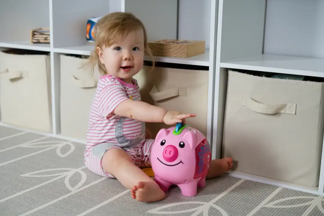 Uśmiechnięte dziecko bawi się różową świnką-skarbonką na tle regału z zabawkami.