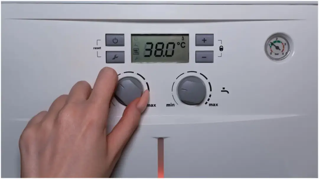 Dłoń reguluje pokrętłem temperaturę na panelu bojlera z cyfrowym wyświetlaczem 38.0°C.