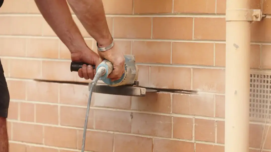 Ręce trzymają szlifierkę kątową, która przecina ceglaną ścianę.