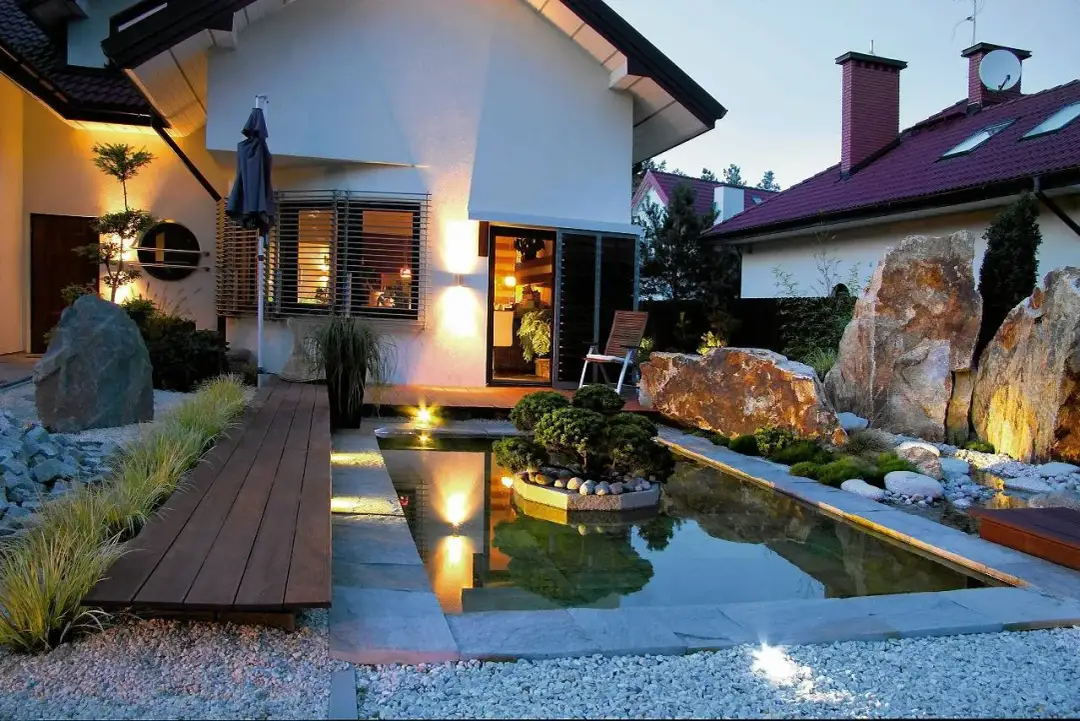 Oświetlenie zewnętrzne domu jednorodzinnego tworzy magiczną atmosferę wokół oczka wodnego i skalnych formacji.