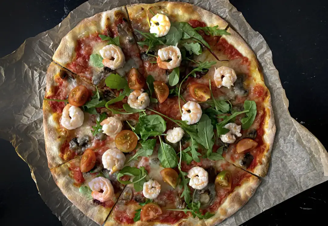 Pyszna pizza z krewetkami, pomidorkami i rukolą, podana na papierze do pieczenia.