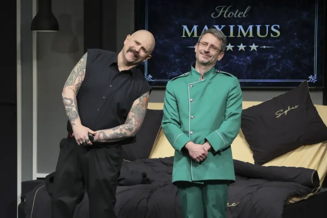 Dwóch aktorów na scenie, jeden w czarnej koszuli z tatuażami, drugi w zielonym mundurze. Tło sugeruje hotel. Spektakl "Rubinowe gody" trwa.