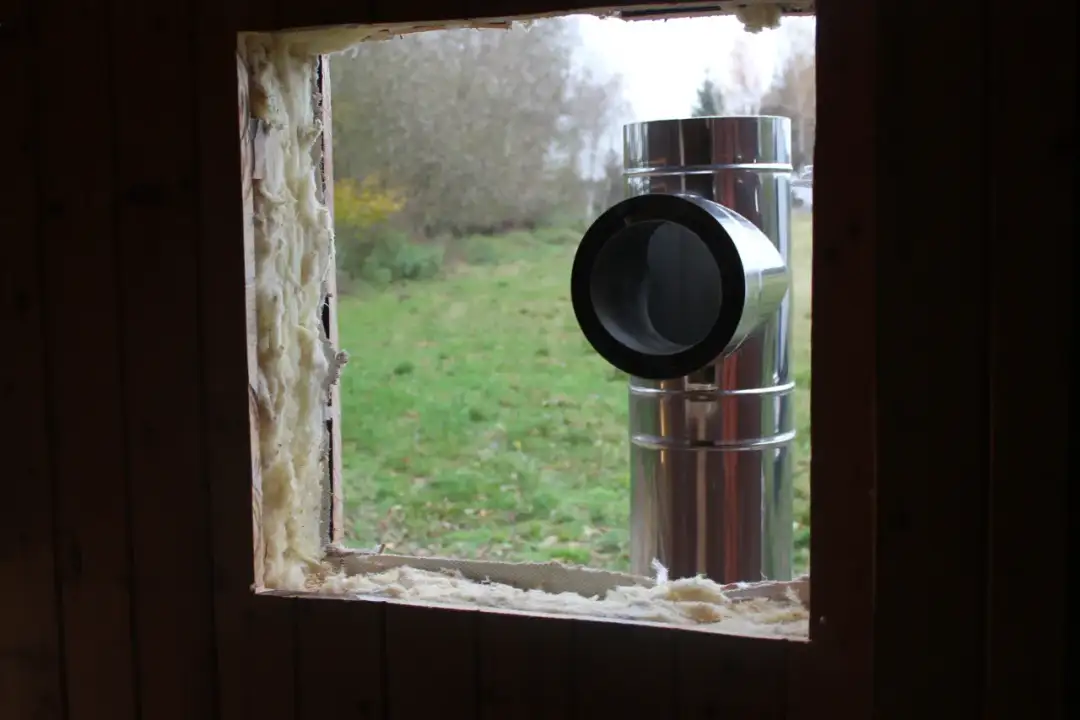 Przejście komina przez ścianę drewnianą, z widocznym izolacyjnym materiałem.