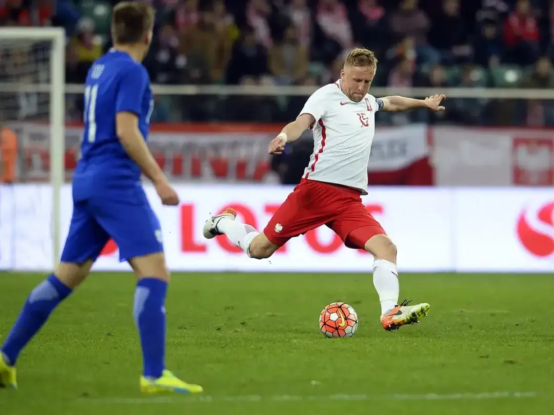 Piłkarz w białej koszulce i czerwonych spodenkach kopie piłkę na boisku.