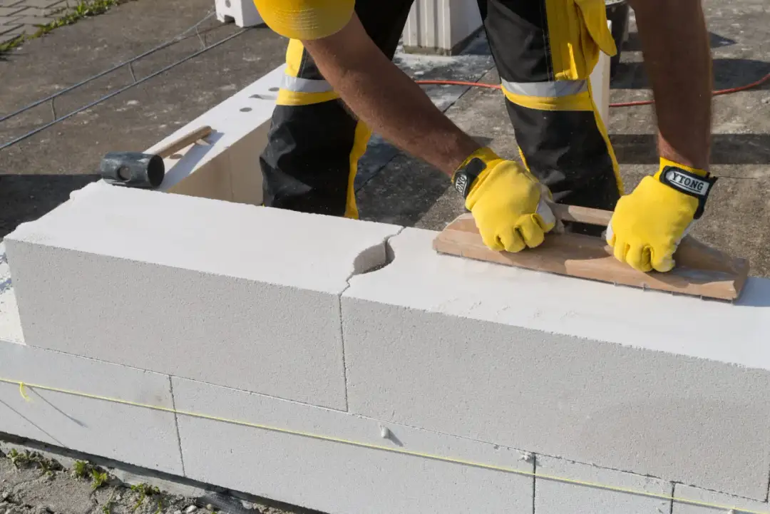 Budowniczy w żółtych rękawicach układa bloczki na fundament. Widać młotek i narzędzie do wygładzania zaprawy.