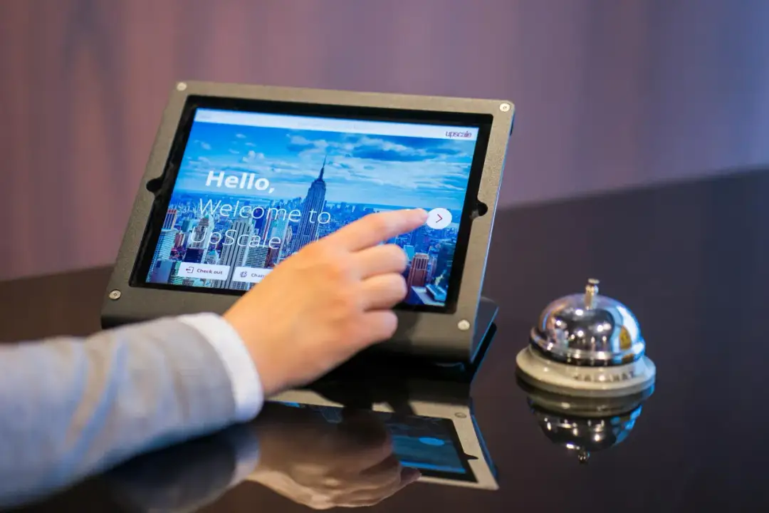 Dłoń dotyka ekranu tabletu z widokiem na Nowy Jork, gdzie można sprawdzić, ile trwa doba hotelowa. Obok stoi dzwonek hotelowy.