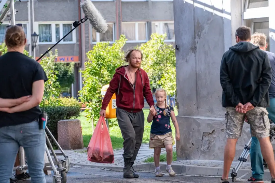 Mężczyzna z dziewczynką idą ulicą, za nimi widać ekipę filmową i mikrofon.