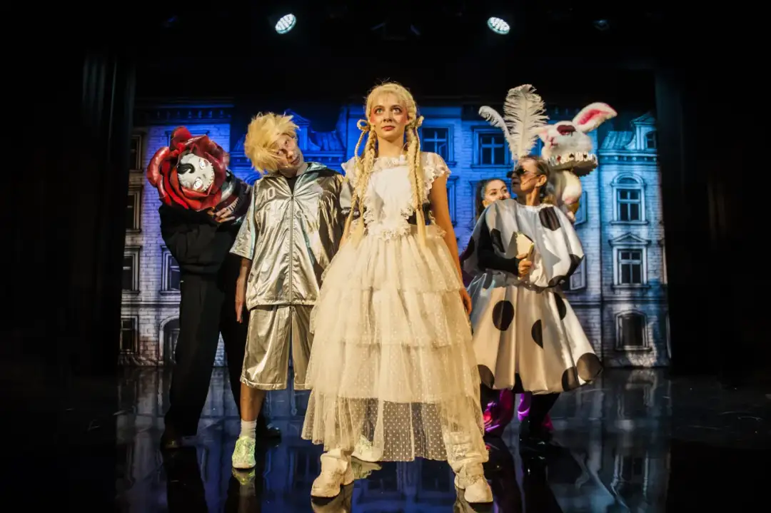 Teatr bajka repertuar dla dzieci: postacie w fantazyjnych strojach, w tym królik i dziewczynka w białej sukni, na tle scenografii.