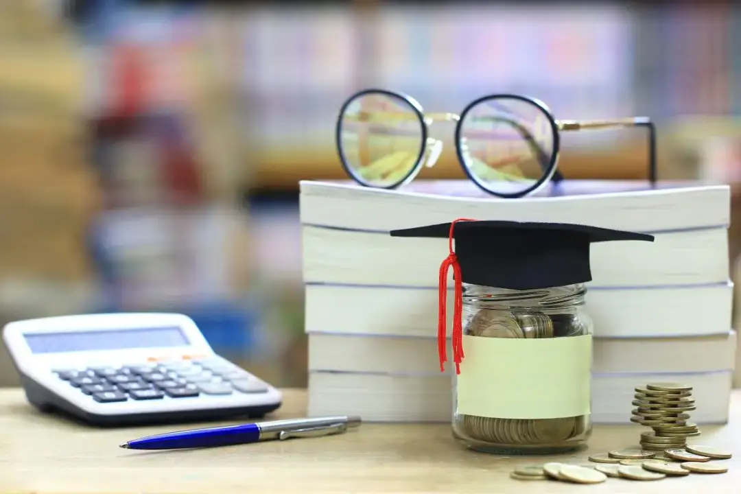 Skarbonka w birecie, stos książek, okulary, kalkulator i długopis. Idealne do zatrudnienia ucznia i studenta do 26 roku życia, brak PIT i ZUS.