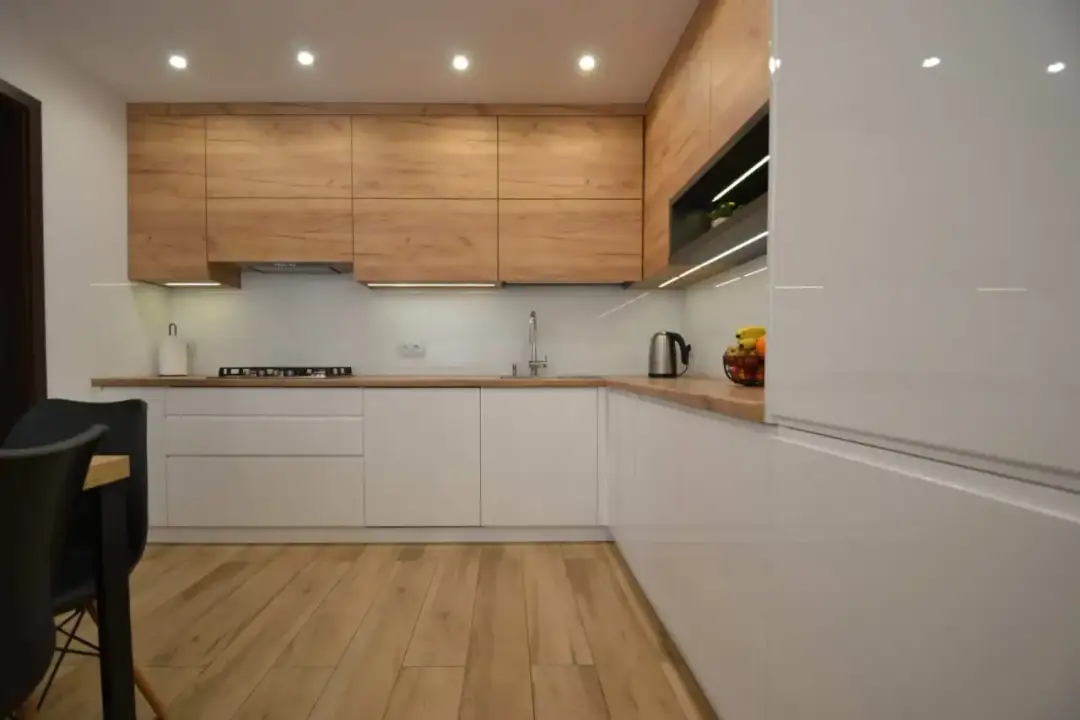 Nowoczesna kuchnia z białymi frontami, drewnianymi szafkami wiszącymi i drewnianą podłogą.