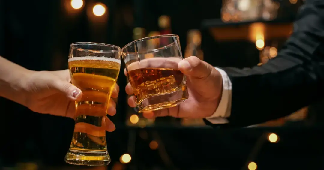 Dwie dłonie unoszą kieliszki: jeden z piwem, drugi z bursztynowym trunkiem. Co lepsze piwo czy wódka? Decyzja należy do Ciebie.