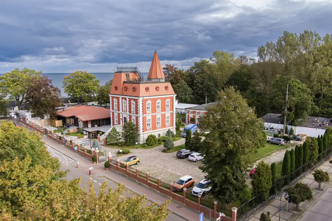 Czerwona willa z wieżyczką i restauracja z pomarańczowym dachem, gdzie można dobrze zjeść w Ustce. Wokół drzewa i parking z samochodami.