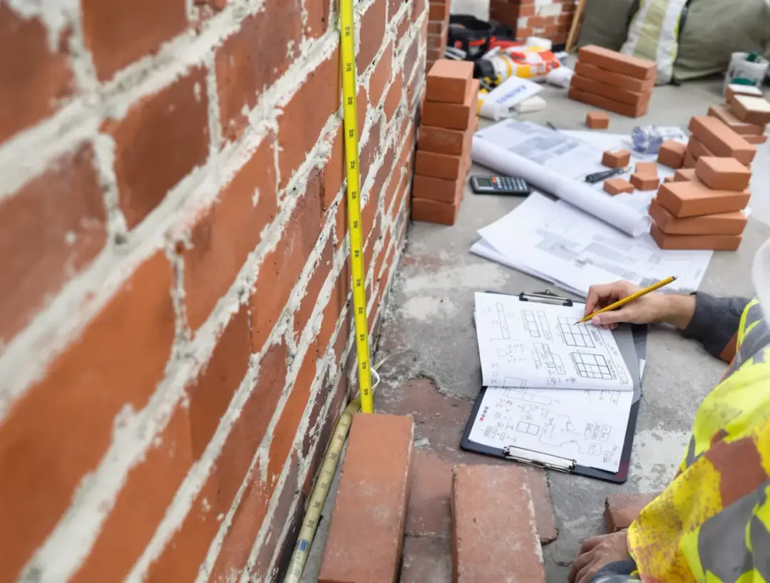 Budowniczy mierzy ścianę, by obliczyć, ile cegieł wchodzi na metr kwadratowy. Na ziemi leżą plany i materiały.