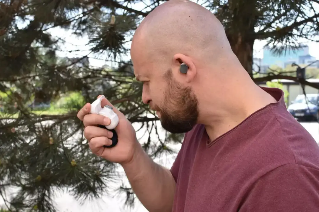 Mężczyzna z zarostem trzyma białe etui z słuchawkami, zastanawiając się, jak zresetować słuchawki bezprzewodowe. Jedna słuchawka jest już w jego uchu.