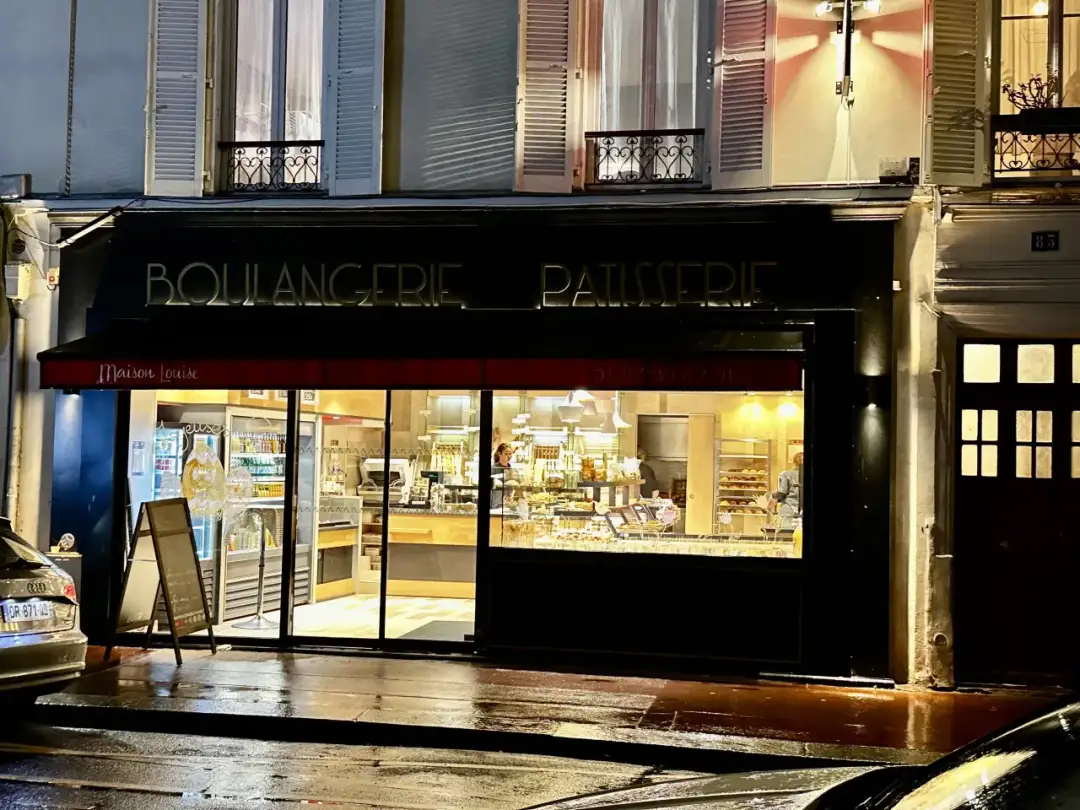 Paryska piekarnia "Maison Louise" nocą. W witrynie widać ciasta i pieczywo. Zastanawiasz się, ile kosztuje obiad w Paryżu?