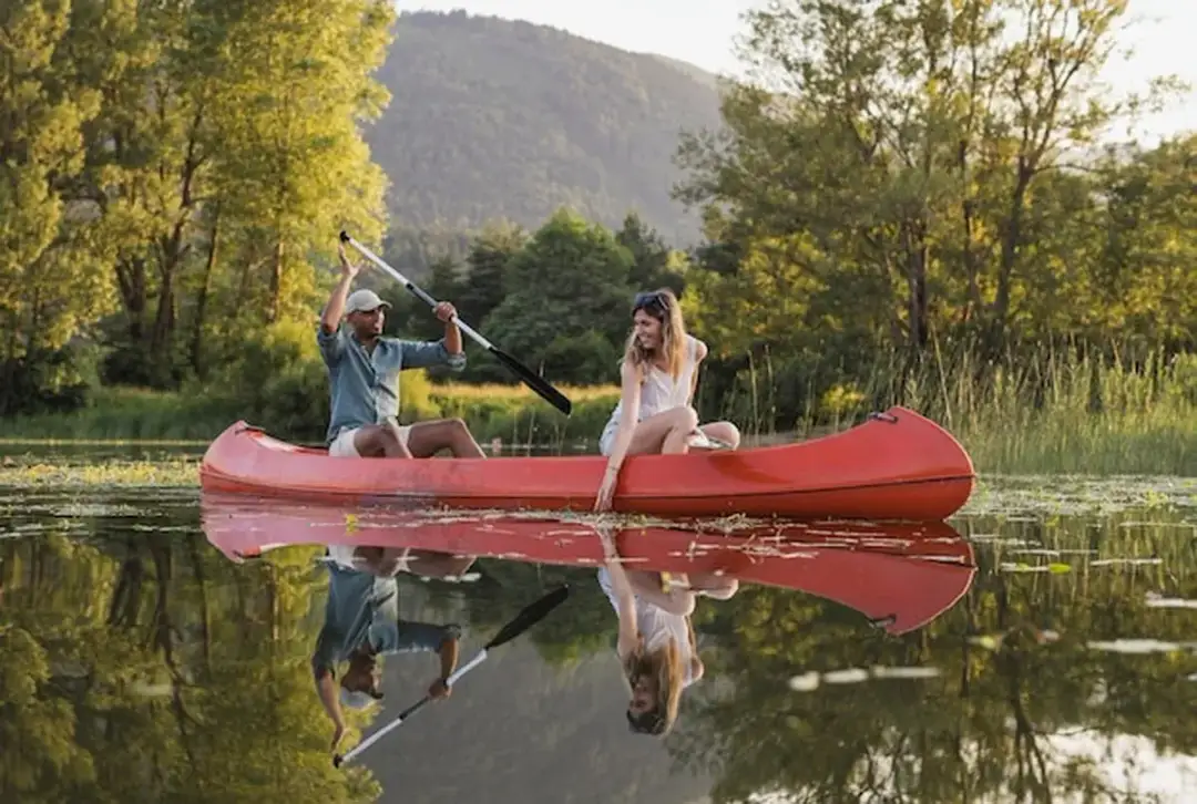 Para płynie kajakiem po jeziorze, otoczona zielenią.