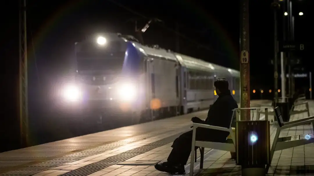 Osoba siedzi na ławce na peronie nocą, zbliża się pociąg z zapalonymi światłami.