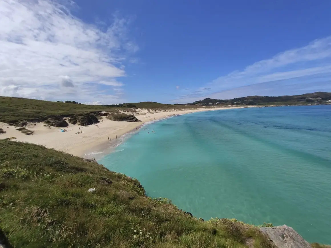 Vista panorámica de la playa de Santa Comba, con arena dorada, aguas turquesas y colinas verdes. Algunas personas disfrutan del sol y el mar.