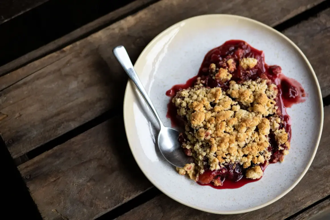 Ein Teller mit warmem Pflaumen Crumble, dazu ein Löffel auf rustikalen Holzdielen.