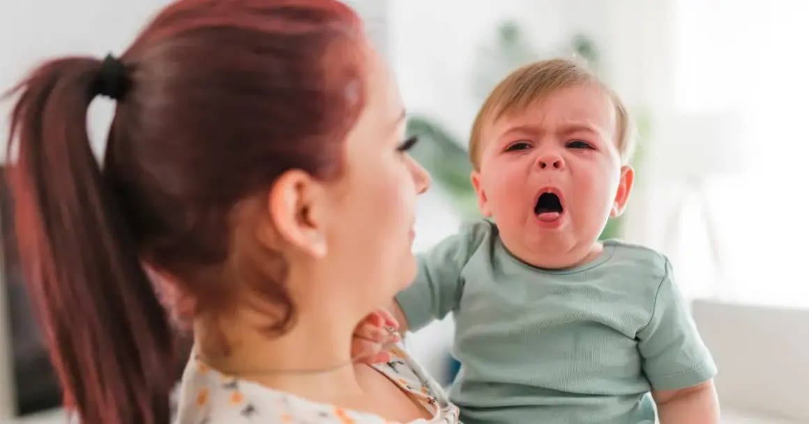 Mama z rudymi włosami tuli płaczące dziecko. Może to być objaw RSV u malucha, który potrzebuje troski.