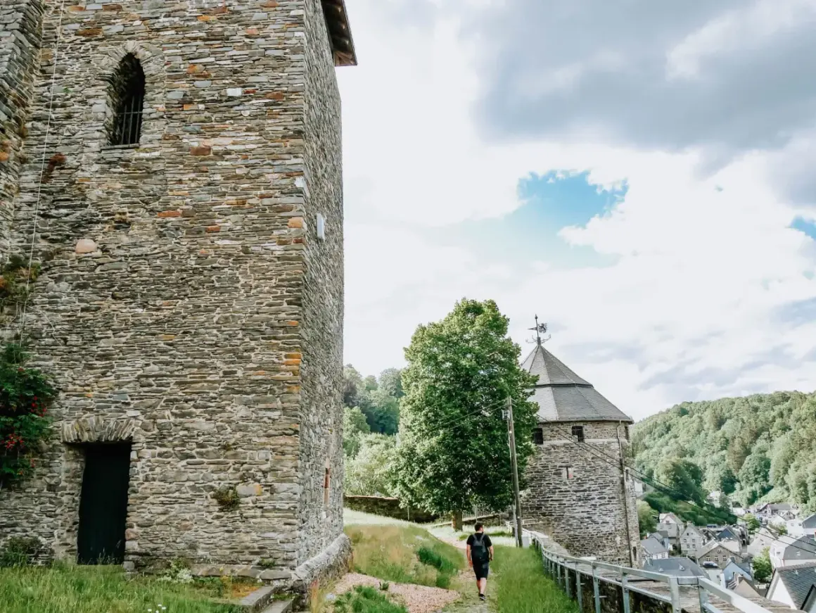 Ein Mann spaziert auf einem Weg zur Burg Monschau, vorbei an alten Steintürmen und grünen Hügeln.