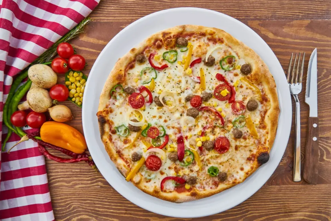 Apetyczna pizza z warzywami i mięsem na talerzu, obok sztućce i składniki.