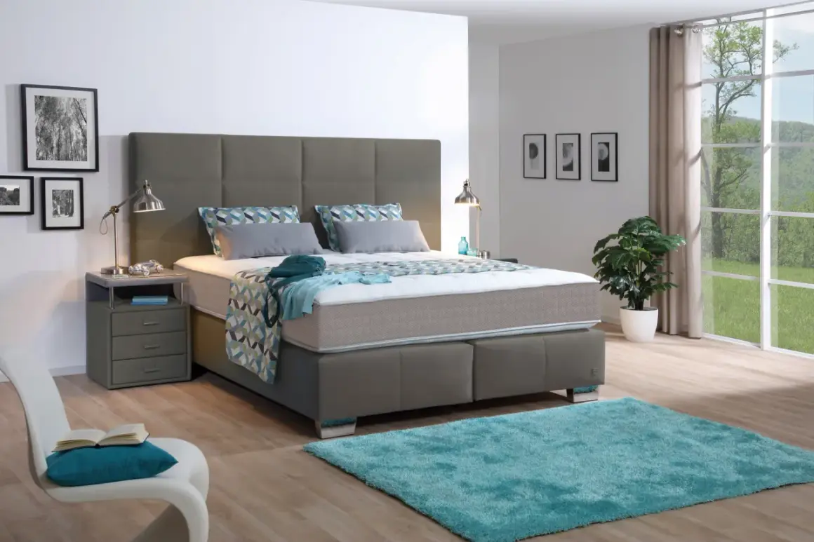 Ein modernes Schlafzimmer mit einem grauen Boxspringbett, das mit einer geometrisch gemusterten Decke und Kissen bezogen ist. Ein türkiser Teppich liegt davor.