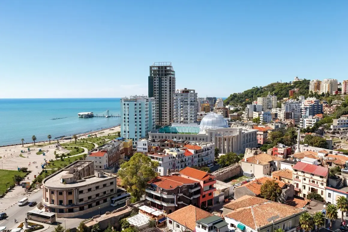 Widok na miasto Durres z plażą w tle. Widać morze, promenadę, budynki mieszkalne i hotelowe oraz wzgórze porośnięte zielenią.