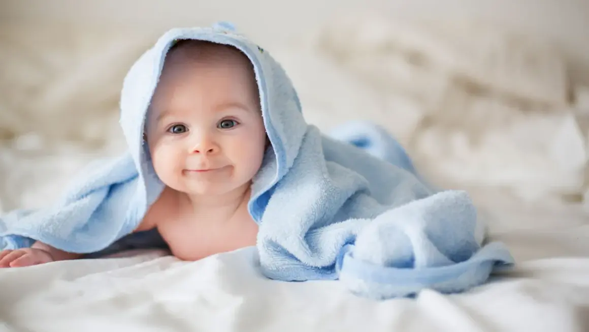 Ein lächelndes neugeborenes Baby liegt auf einem weißen Bett, eingewickelt in ein hellblaues Handtuch.