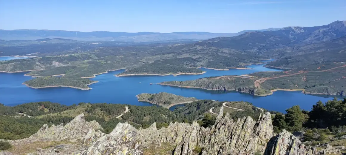 Vista panorámica del embalse de Puentes Viejas, con sus aguas azules serpenteando entre colinas verdes y montañas al fondo.