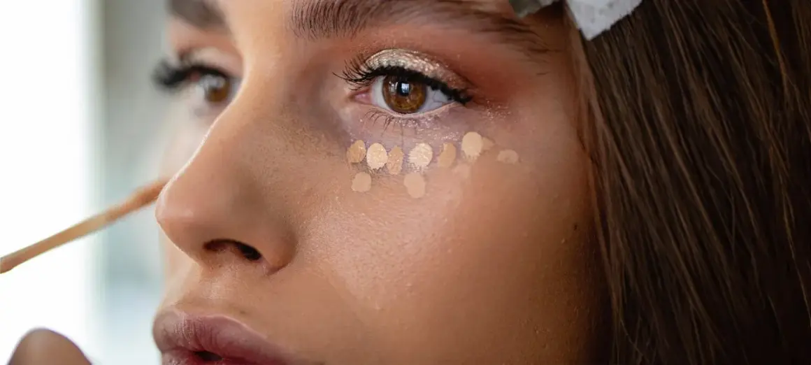 Nahaufnahme einer Frau, die Concealer unter den Augen aufträgt. Mehrere helle Tupfer bilden eine Reihe, bereit zum Verblenden.