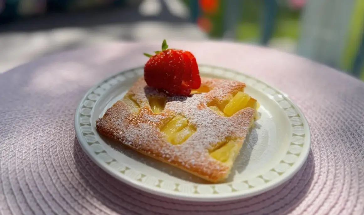 En bit saftig ananaskaka i långpanna, pudrad med florsocker och toppad med en färsk jordgubbe. Perfekt för en sommardag.