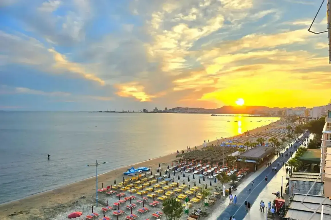 Zachód słońca nad Durrës plaża. Rzędy leżaków i parasoli czekają na plażowiczów, a w oddali widać panoramę miasta.